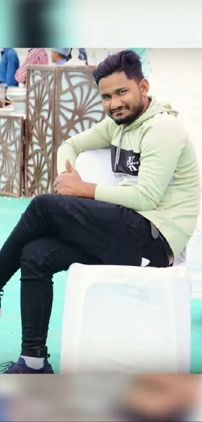 Man in casual attire posing on a white chair against a green background.