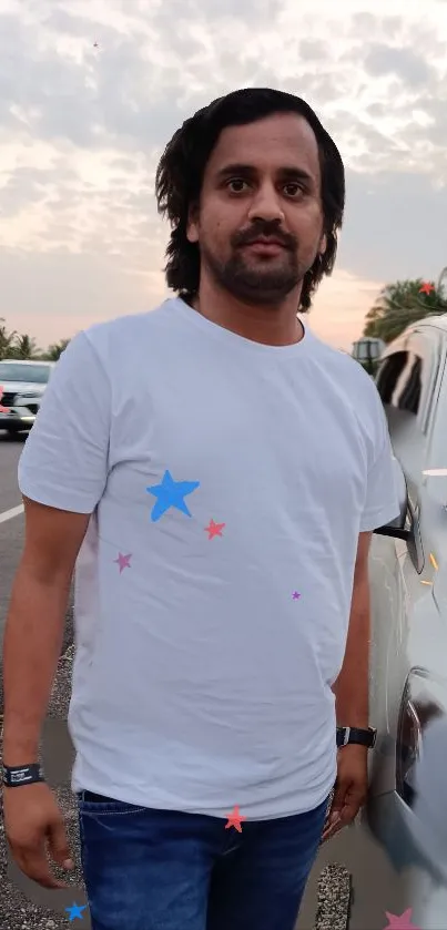 Man in white t-shirt standing by road with cars and sky.