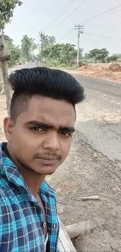 Casual selfie with a serene roadside background and blue shirt.