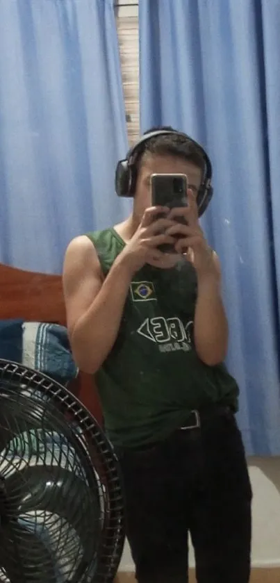 Casual selfie in a mirror with headphones, featuring blue curtains and a fan.