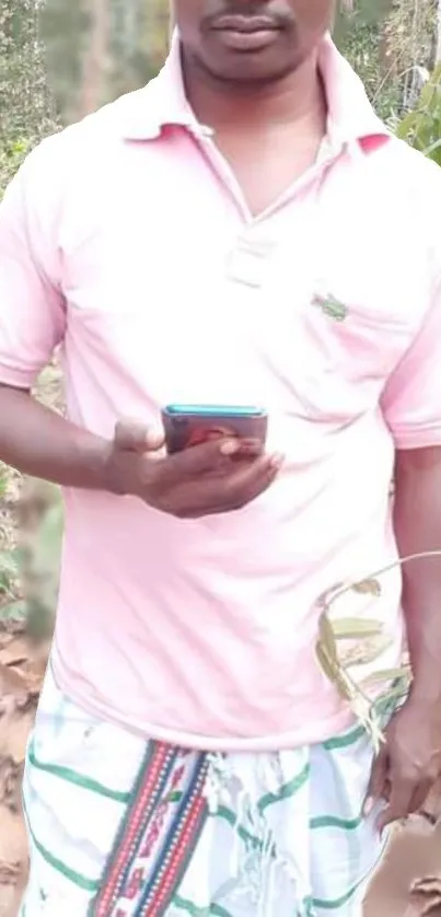 Man in pink shirt standing outdoors, holding a phone in a natural setting.