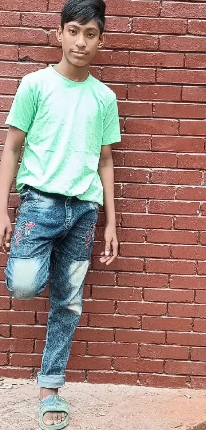 Teen boy leaning against a red brick wall in casual attire.