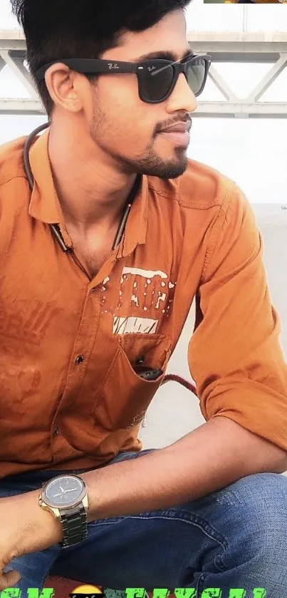 Young man in sunglasses with burnt orange shirt and watch, seated outdoors.