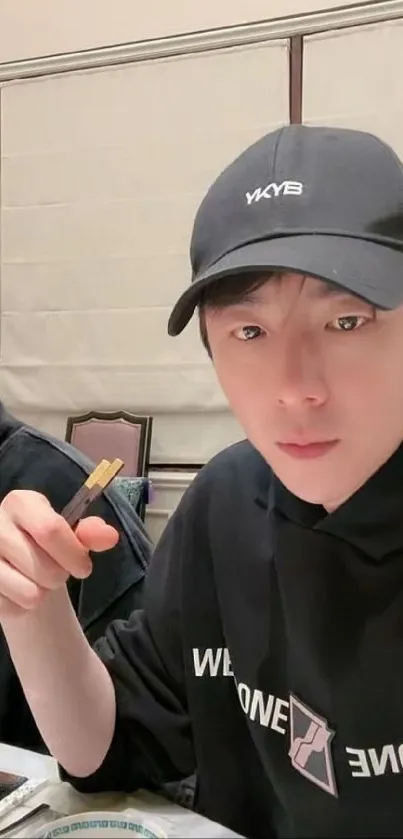 Young man dining with chopsticks, wearing a casual cap and hoodie.