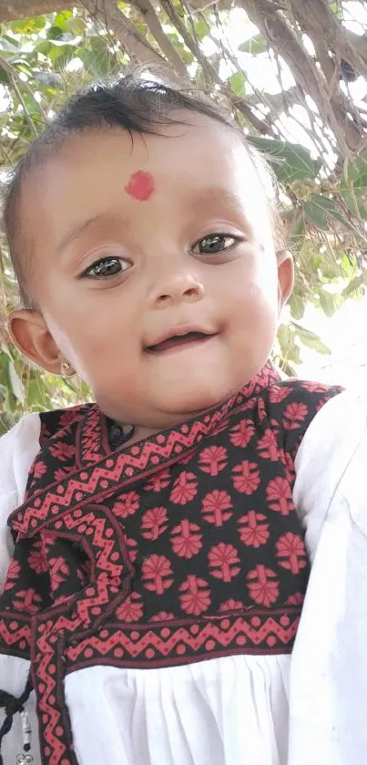 Baby in traditional attire under a tree with a charming expression.