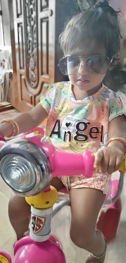 Child with sunglasses on pink bike indoors.