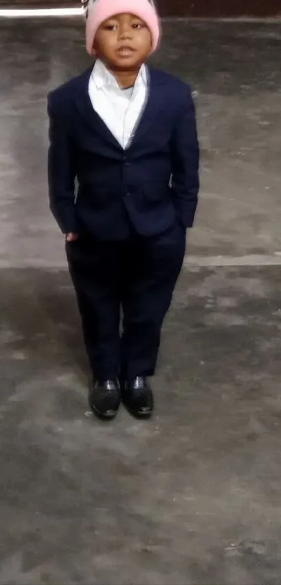 Child in a formal suit and pink hat on a gray floor.