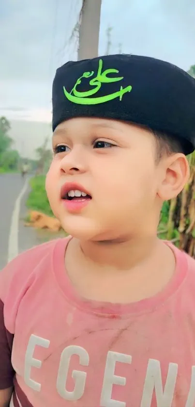 A child wearing a black hat, standing outdoors with greenery in the background.