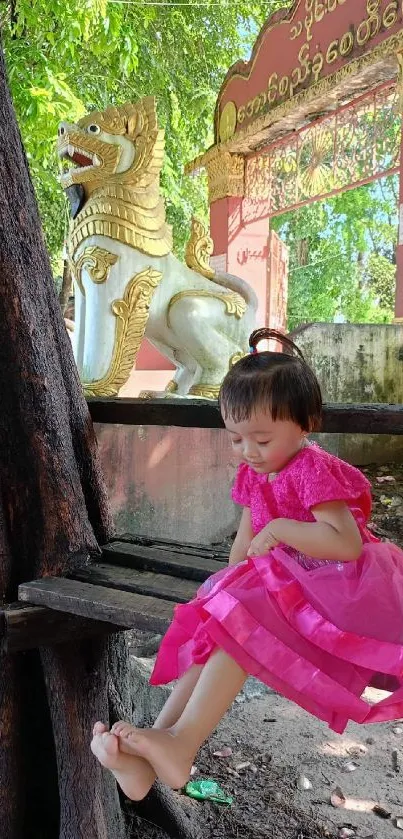 Young girl in pink dress sitting by a tree with cultural backdrop.