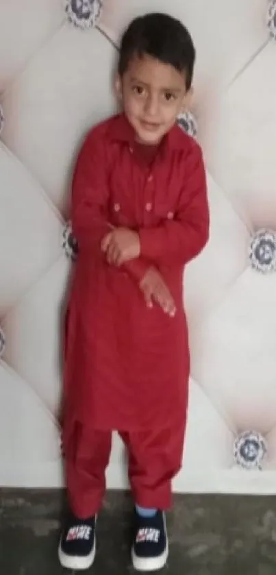 Smiling child in red traditional attire standing against a decorative wall.