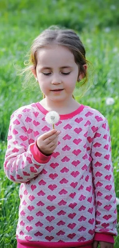 Young girl in pink sweater holding dandelion in green field.