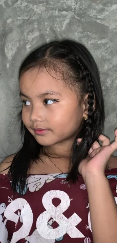 Portrait of a young girl with braided hair against a gray textured wall background.