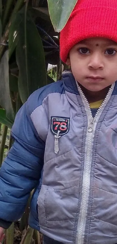 Child in a red hat standing outdoors amid green plants.