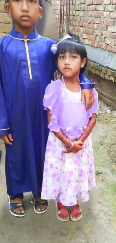 Two children in traditional outfits standing by a brick wall.