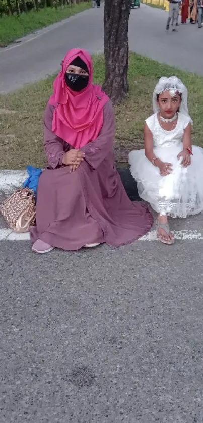 Two people sitting outdoors, a woman in pink and a girl in white, on a serene street.