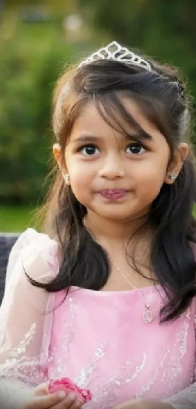 A young girl with a tiara, dressed in pink, smiling outdoors.