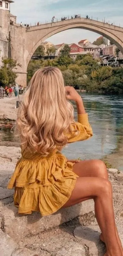 Woman in yellow dress sitting near a river under a stone bridge.