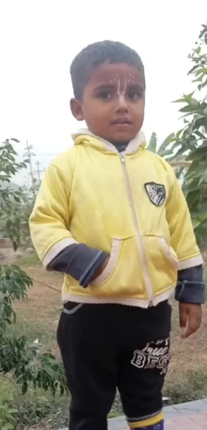 Toddler in yellow hoodie outdoors with greenery background.