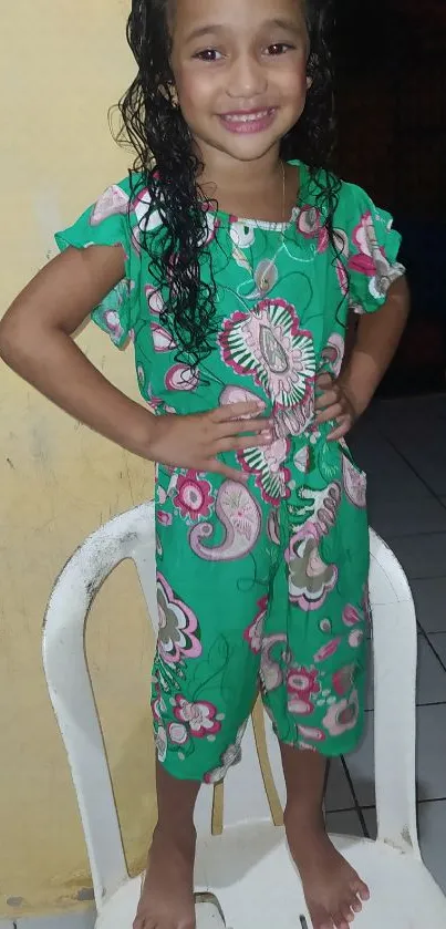 Smiling child in vibrant green floral outfit sitting on a chair.