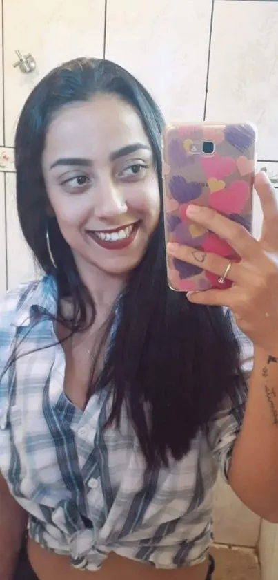 Woman in plaid shirt taking a mirror selfie with a heart-patterned phone case.