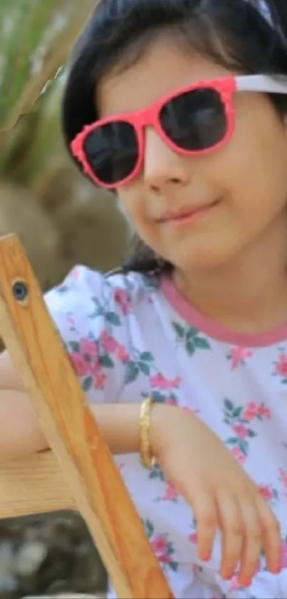 Child wearing pink sunglasses and floral dress on wooden chair.