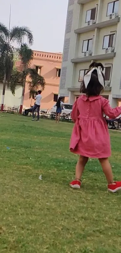 Child in pink dress playing on grass with building in background.