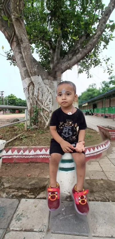 Child sitting on a post in a garden with red shoes and a tree background.