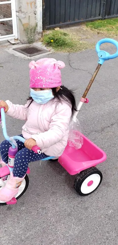 Young child in mask rides pink tricycle.