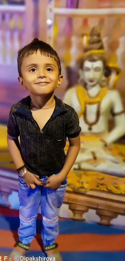 A smiling child stands before a colorful background with a traditional statue.