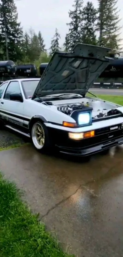 Classic white Toyota AE86 parked on a wet driveway with trees in the background.