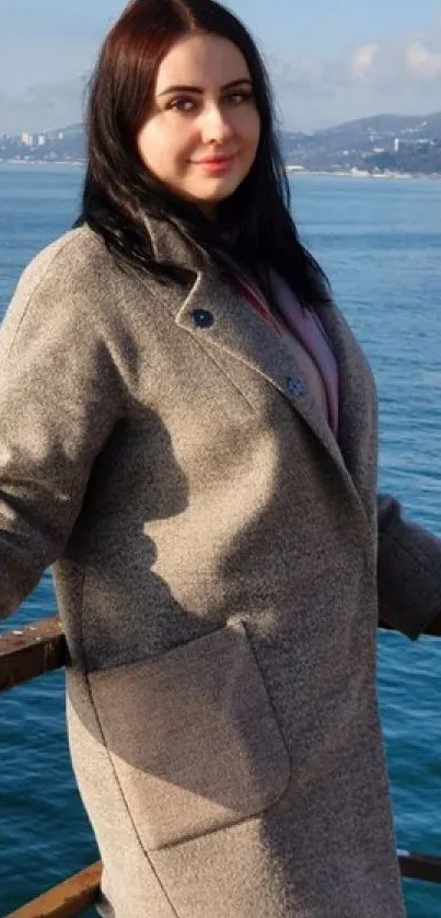 A woman in a coat by a serene blue ocean with distant hills.