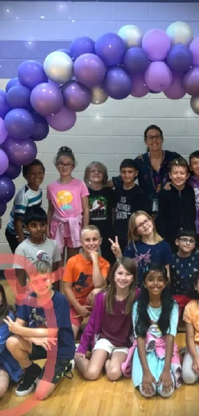 A group of children and teacher under a colorful balloon arch in a gym.