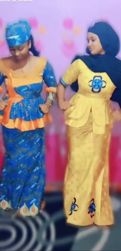 Two women in vibrant traditional attire dancing against a pink backdrop.