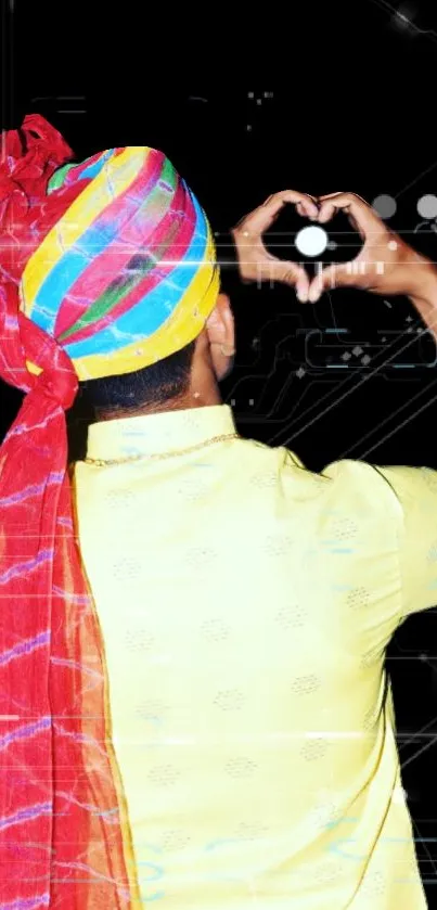 Person in colorful turban making heart shape with hands against black background.