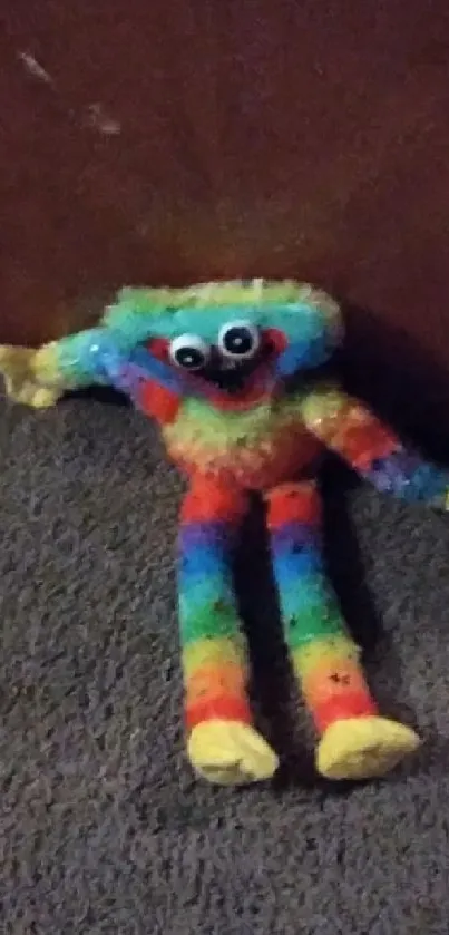 Colorful plush toy leaning against a brown backdrop on gray carpet.