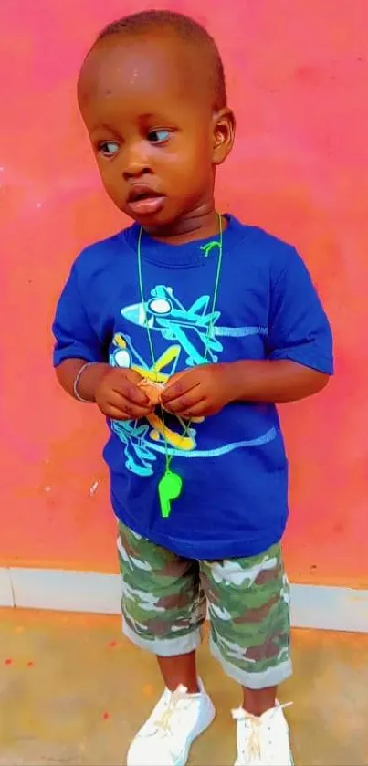 Toddler in blue shirt and camo shorts against coral wall.