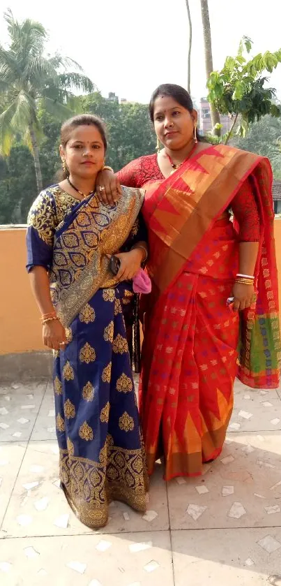 Two women in vibrant sarees with a natural background.