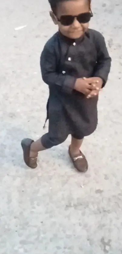 Stylish kid in sunglasses and dark outfit posing confidently on pavement.