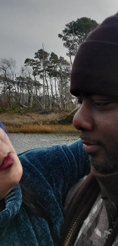 Couple embracing by a calm lake with forest backdrop.