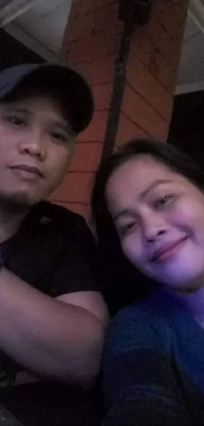 Couple in a cozy evening setting, smiling toward the camera with warm lighting.