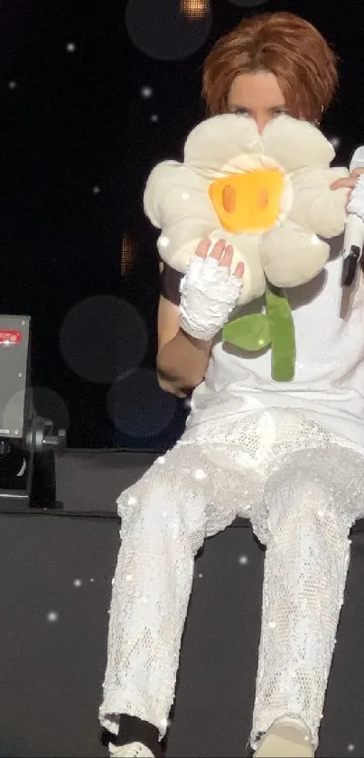 Performer holding a plush flower on stage in cozy setting.