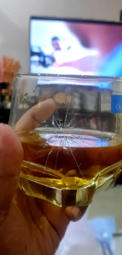 Hand holding cracked whiskey glass with TV background.