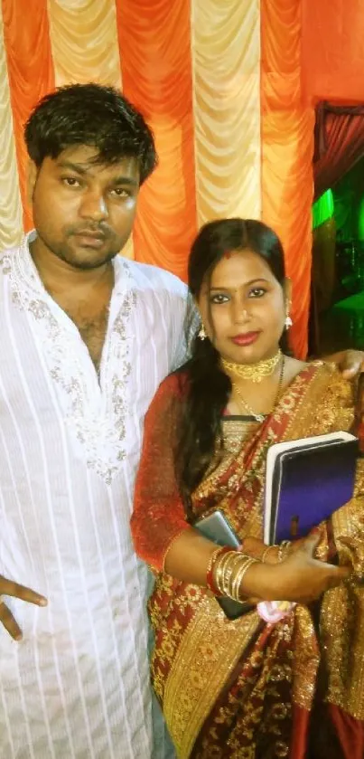 A couple in traditional attire at a vibrant cultural event with orange drapery.