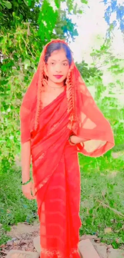 A woman in a vibrant red saree stands gracefully in a lush green outdoor setting.