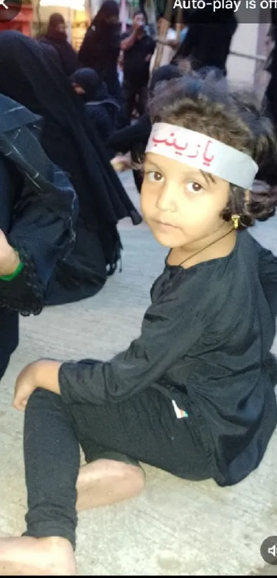 Child in traditional gathering, sitting on the ground.