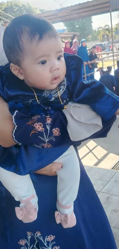 Cute baby in a blue embroidered dress being held outdoors.