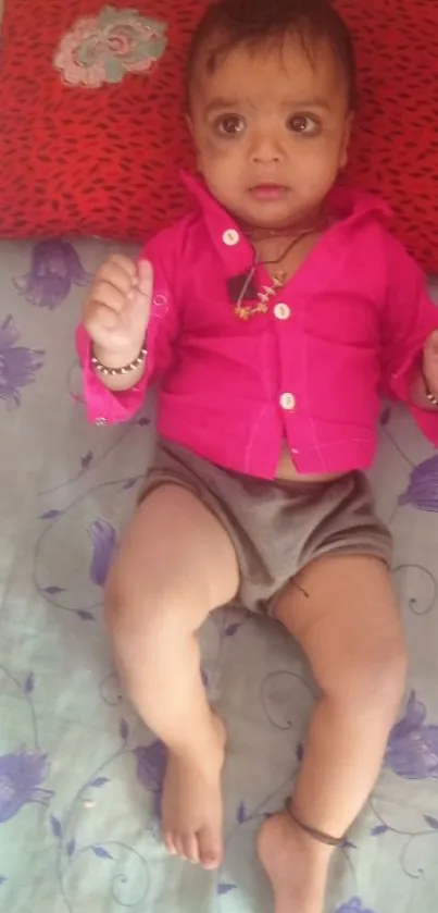 A cute baby in pink shirt and grey shorts on floral bedding.