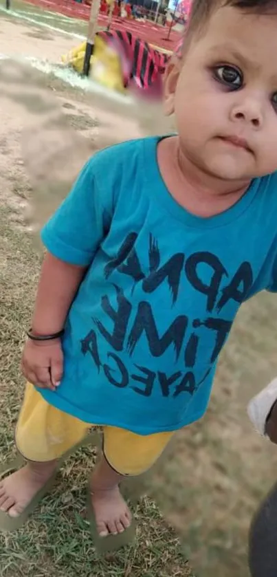 Cute baby in blue T-shirt and yellow shorts standing outdoors.