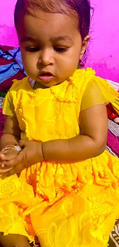 Cute baby in a vibrant yellow dress sitting on a colorful bedspread.