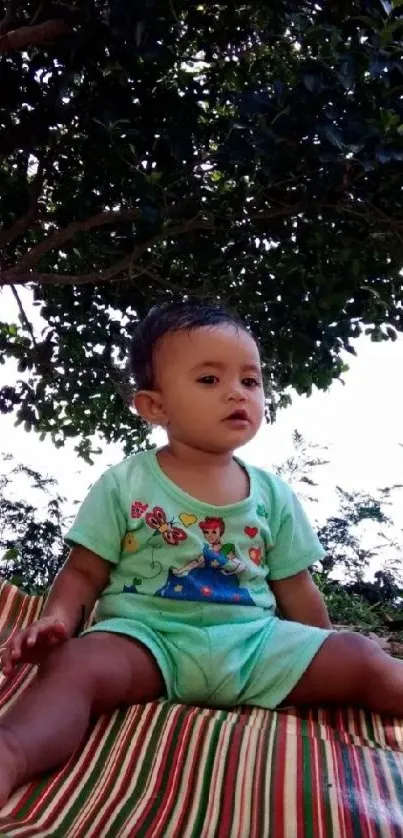 Adorable baby sits outdoors on a colorful mat under lush green trees.